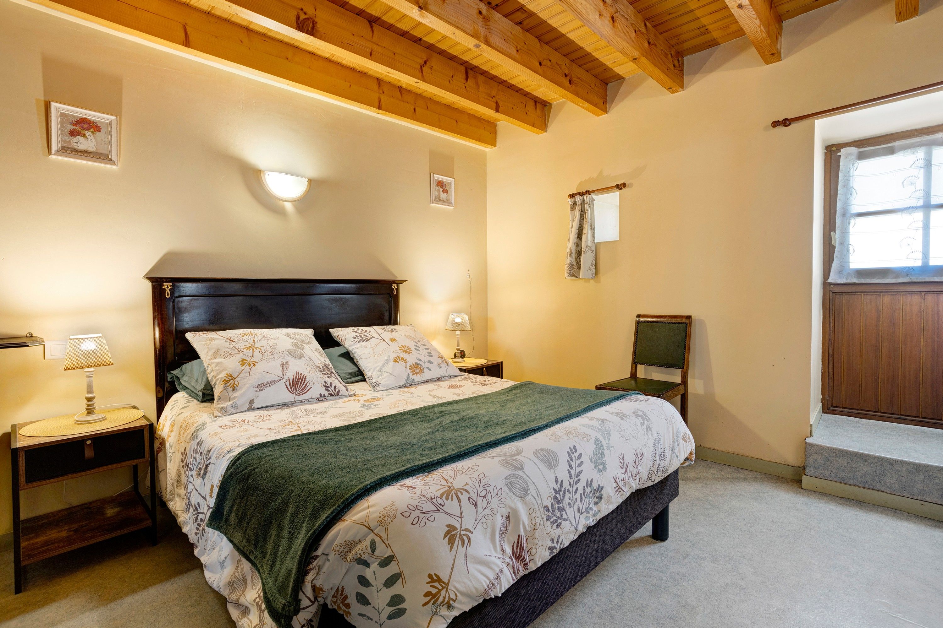Une chambre avec un plafond en bois, un lit recouvert d’une literie à motifs floraux, une table de chevet avec une lampe et une fenêtre avec un rideau.