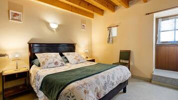 Une chambre avec un plafond en bois, un lit recouvert d’une literie à motifs floraux, une table de chevet avec une lampe et une fenêtre avec un rideau.