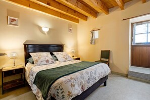 Une chambre avec un plafond en bois, un lit recouvert d’une literie à motifs floraux, une table de chevet avec une lampe et une fenêtre avec un rideau.
