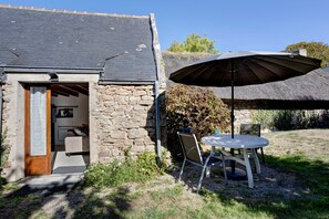Un bâtiment en pierre avec un toit de chaume, une porte en bois et un petit patio comprenant une table et des chaises.