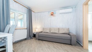 Ein Zimmer mit grauem Sofa, einem weißen Tisch mit Stuhl, einem Fenster mit Vorhängen und einem Heizkörper.