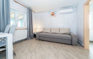 Ein Zimmer mit grauem Sofa, einem weißen Tisch mit Stuhl, einem Fenster mit Vorhängen und einem Heizkörper.