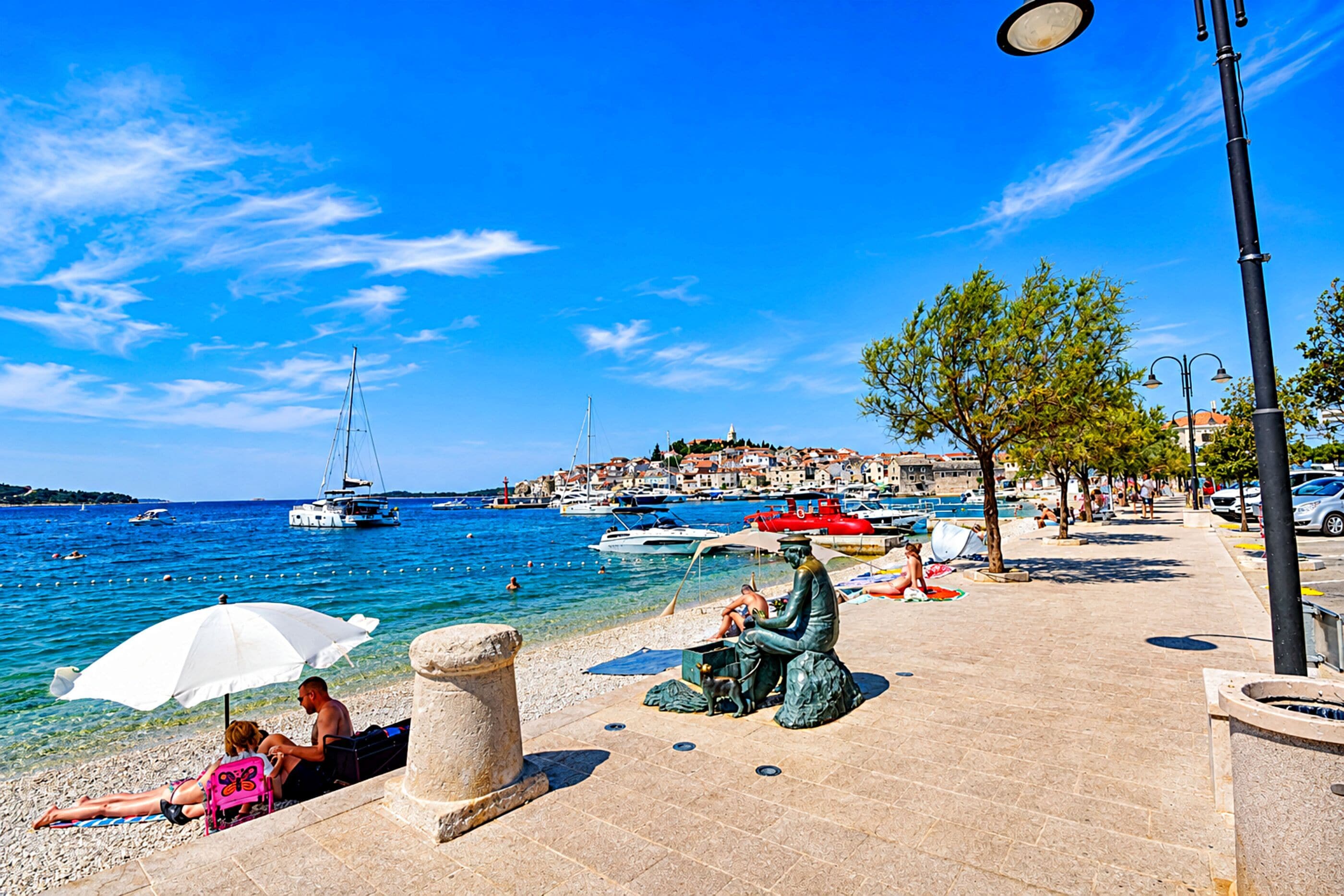 Eine Küstenpromenade mit klarem, blauem Wasser, Booten und einer Statue.