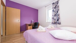 Ein Schlafzimmer mit violetten Wänden, einem Holzschrank, einem Schreibtisch mit Stuhl, einem Bett mit Kissen und Handtüchern sowie einem Fenster mit Vorhängen.
