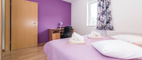 A bedroom with a purple wall, a wooden wardrobe, a desk with a chair, a bed with pillows and towels, and a window with curtains.