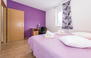 Ein Schlafzimmer mit violetten Wänden, einem Holzschrank, einem Schreibtisch mit Stuhl, einem Bett mit Kissen und Handtüchern sowie einem Fenster mit Vorhängen.