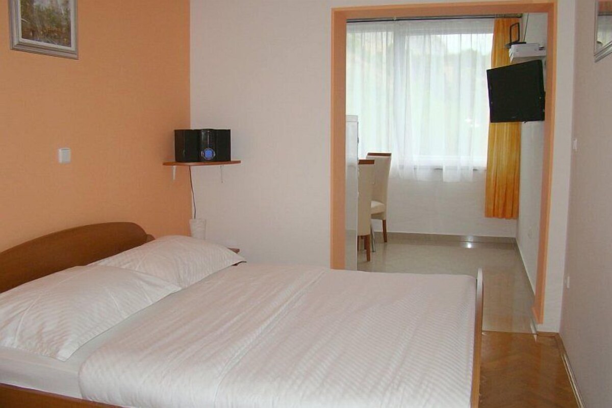 A bedroom with a bed, a TV on a stand, a desk with a chair, and a window with curtains.