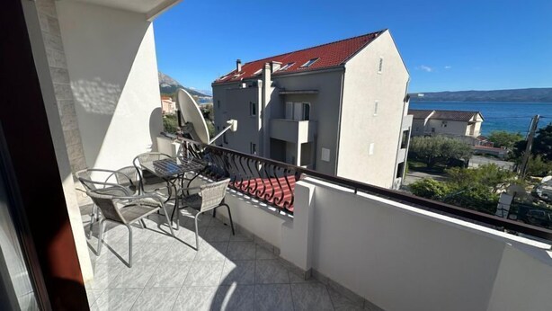 A balcony with a table and chairs, a red awning, and a view of buildings and water. - Apartments Vila Paula (Dugi Rat)