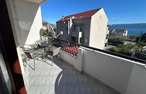 Een balkon met een tafel en stoelen, een rode zonnescherm en uitzicht op gebouwen en water.