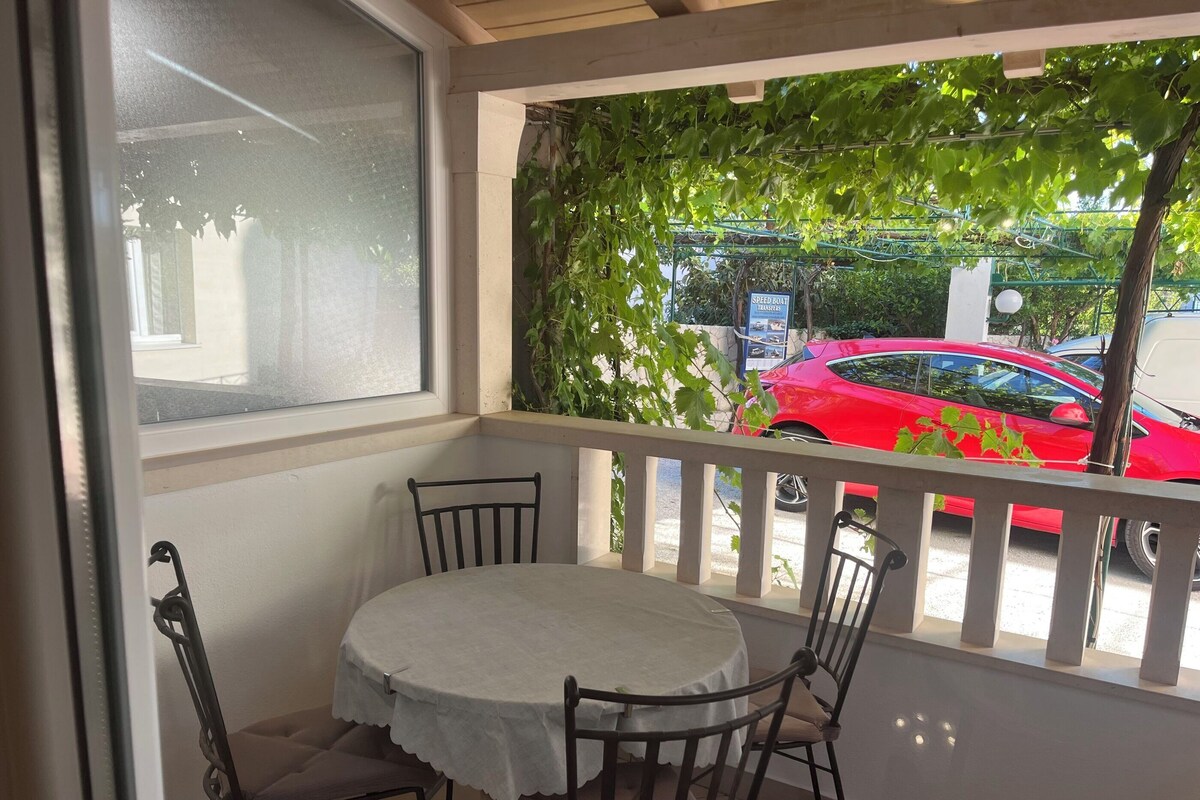A balcony with a table and chairs, a view of a red car, and greenery.