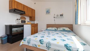 Een compacte kamer met een bed, een kitchenette met een kookplaat, magnetron en kastjes, een kleine prullenbak en een wandplank.