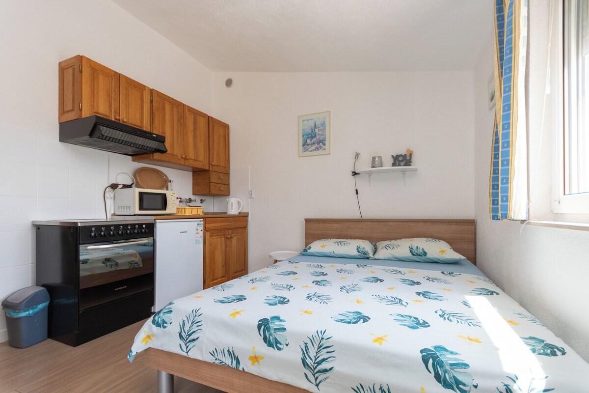 Ein kompaktes Zimmer mit einem Bett, einer Küchenzeile inklusive Herd, Mikrowelle und Schränken, einer kleinen Mülltonne und einem Wandregal.