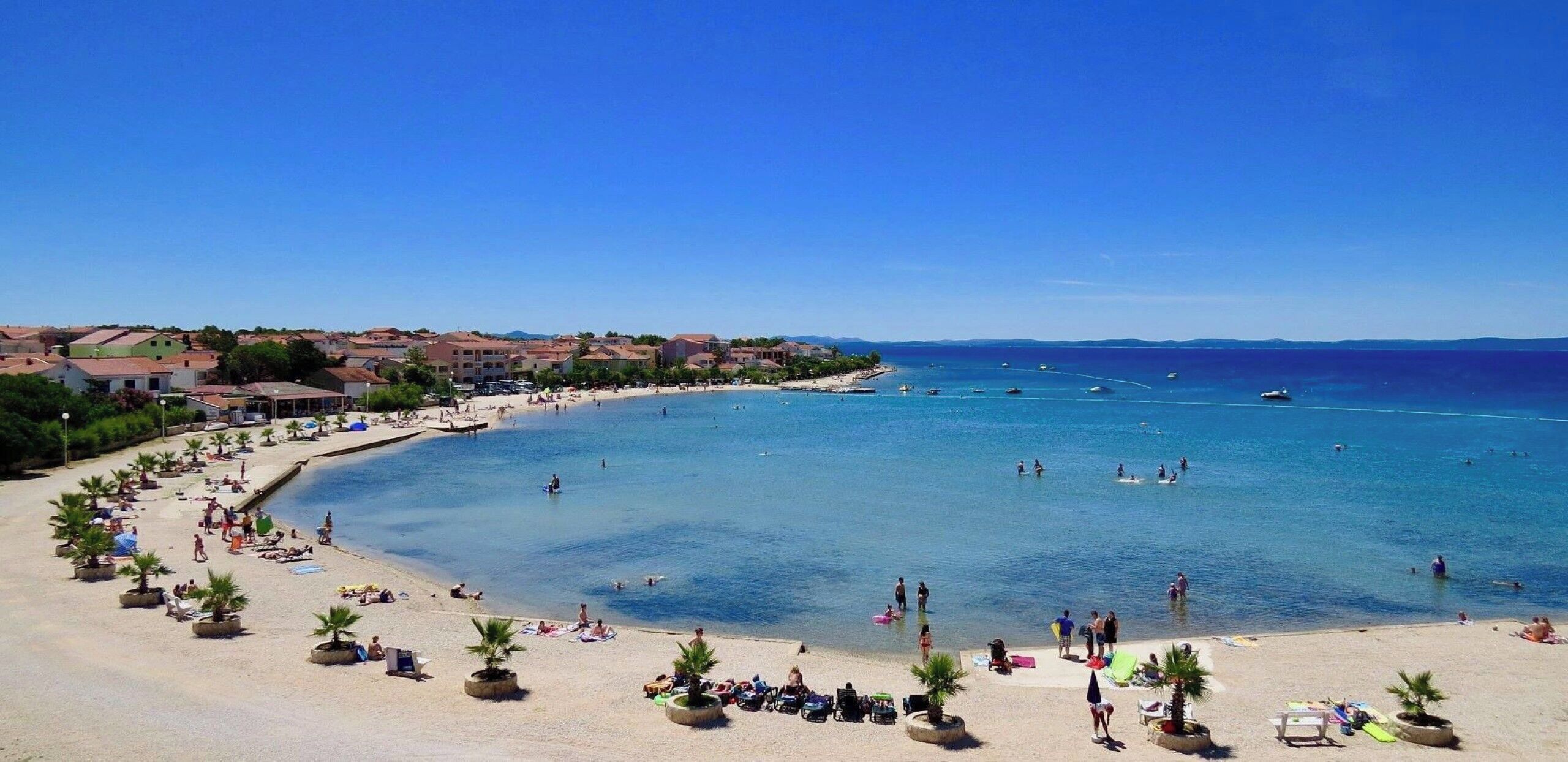 A coastal beach with clear blue water, a sandy shore, and buildings in the background.