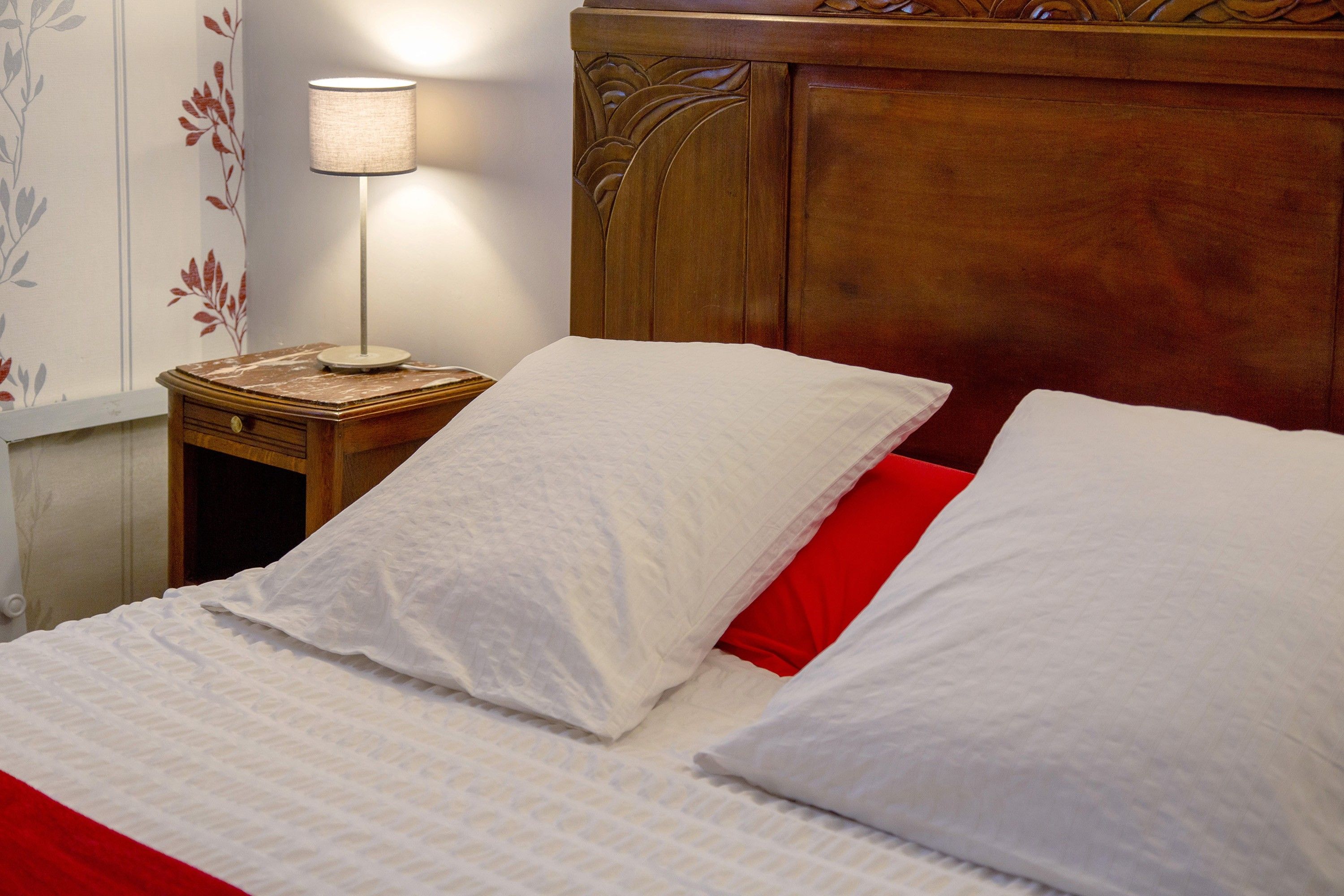 Un lit bien fait, avec du linge de lit blanc et un oreiller rouge, une lampe de chevet et une table de chevet en bois dotée d’un tiroir.