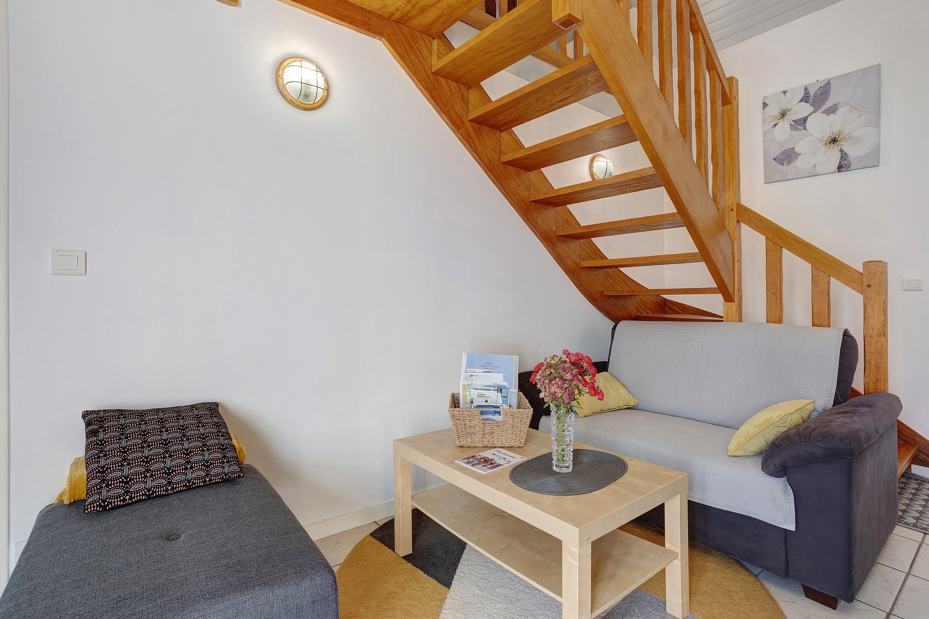 A cozy living area with a wooden staircase, a sofa, a small table with a vase of flowers, and a patterned pillow.