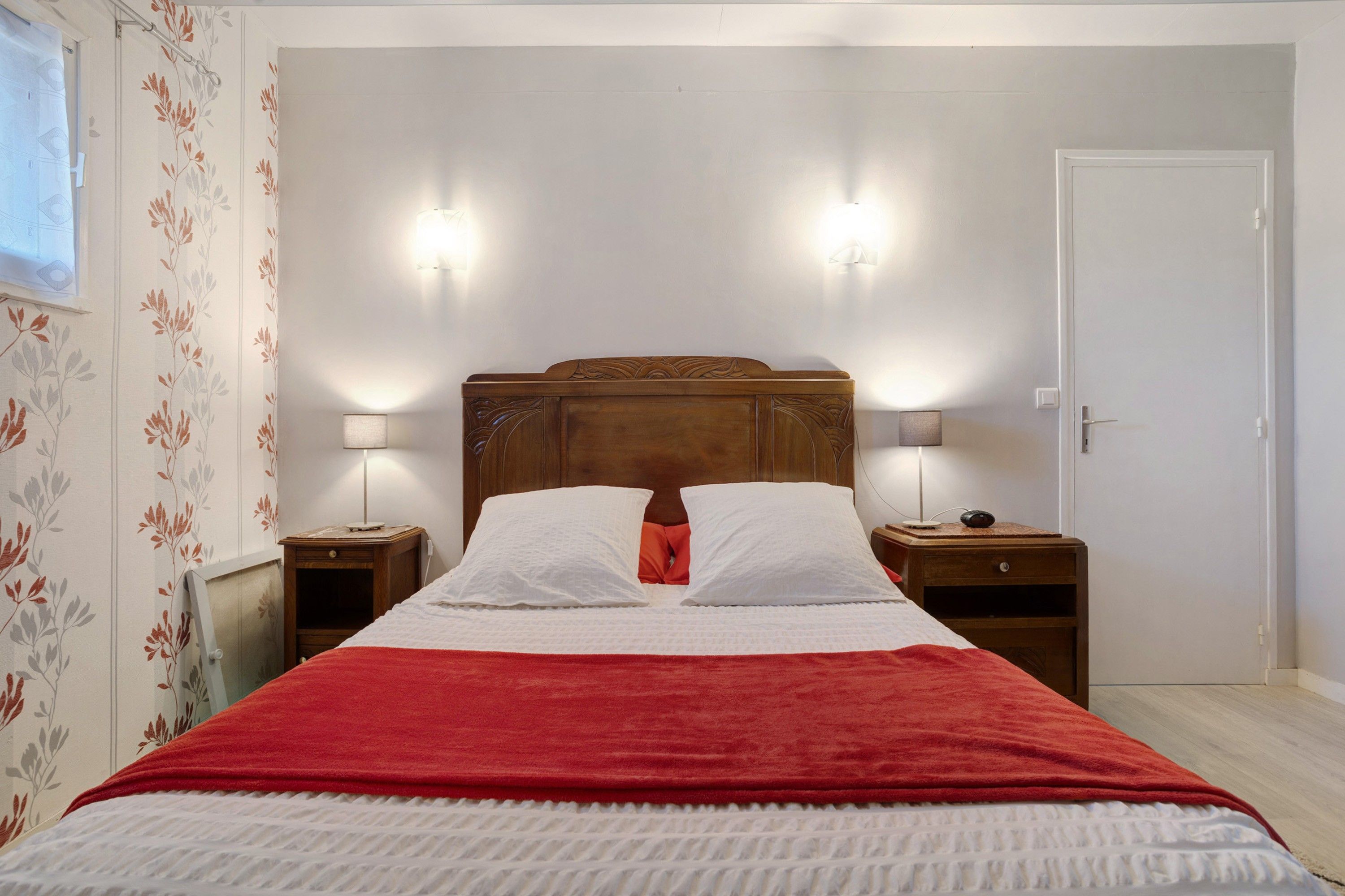 A neatly made bed with a red blanket, two bedside lamps, and a wooden headboard.