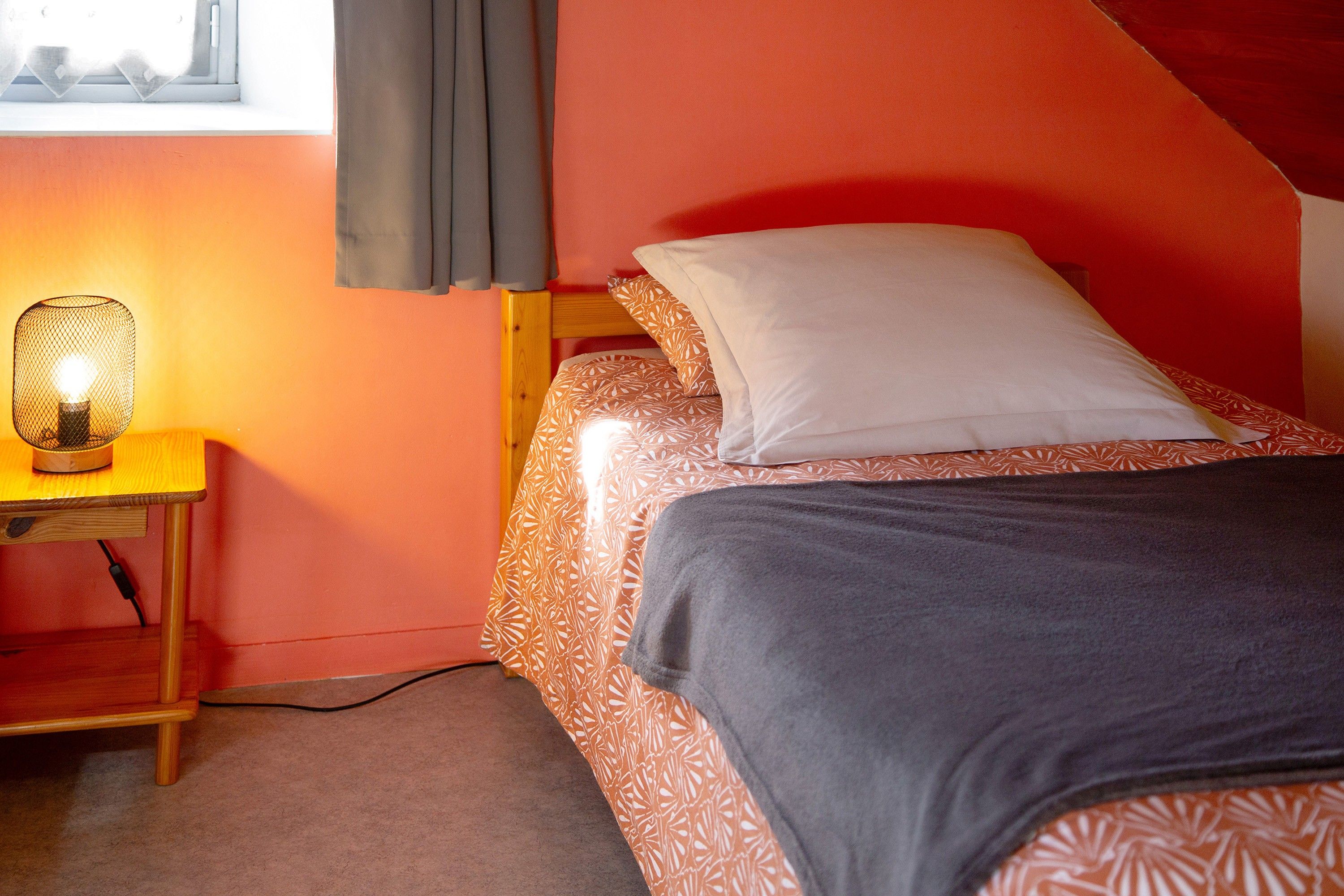 Una cama individual con una colcha estampada, una mesita de noche con lámpara y una ventana con cortinas.