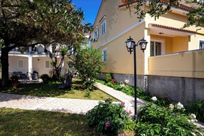 A building with a yellow facade, a stone pathway, and a tree with a bench. - Apartments Mladenka 2 (Rab)