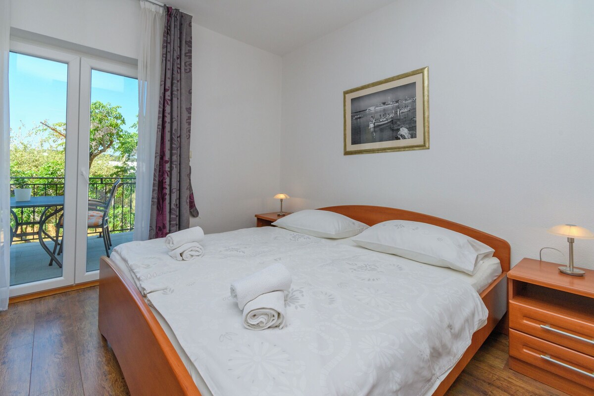 A bedroom with a bed, a nightstand, a framed picture on the wall, and a balcony with a table and chairs.