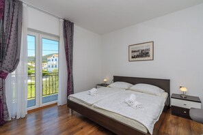 A bedroom with a bed, two bedside tables, a nightstand, a window with curtains, and a framed picture on the wall. - Apartments Mladenka 2 (Rab)