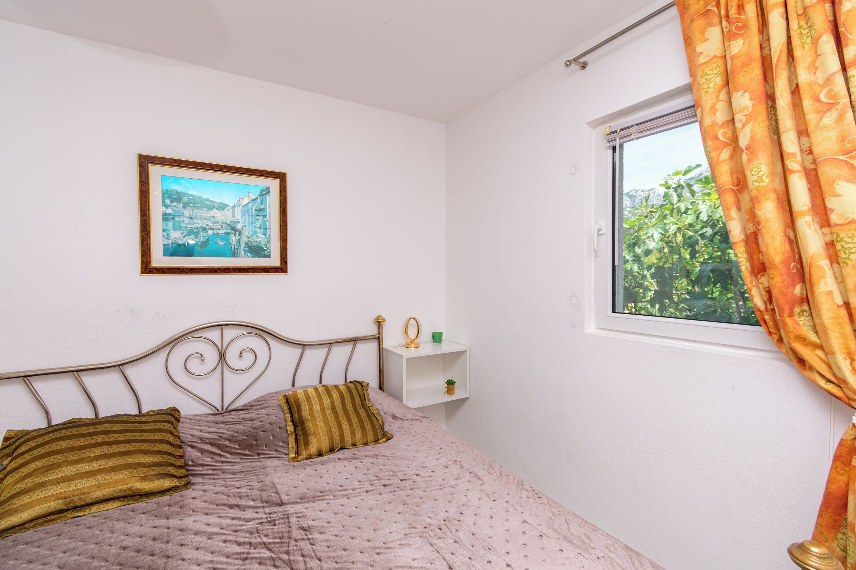Una cama con cabecera, un cuadro enmarcado en la pared, un estante con objetos y una ventana con cortinas.