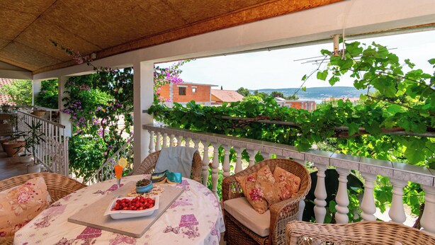 Un espacio de estar al aire libre cubierto, con mobiliario de mimbre, una mesa con mantel floreado y vistas a zonas verdes y edificios al fondo.