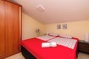 Un dormitorio con una colcha roja, un armario de madera, una mesita de noche con una lámpara y un ventilador fijado a la pared.