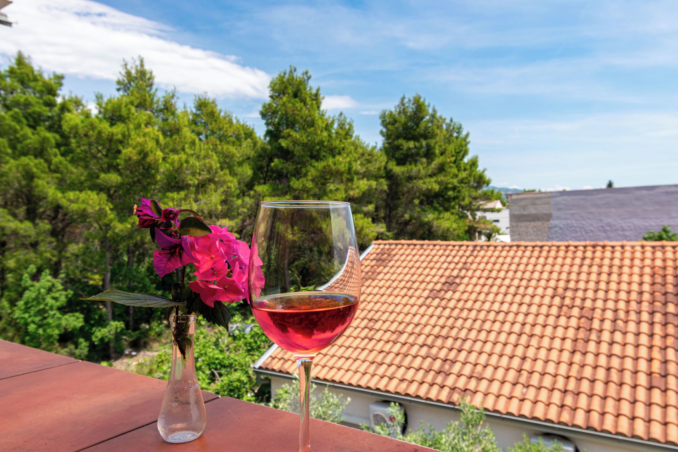 Ein Glas Rotwein und eine Vase mit rosa Blumen auf einem Holztisch, im Hintergrund ein Ziegeldach.