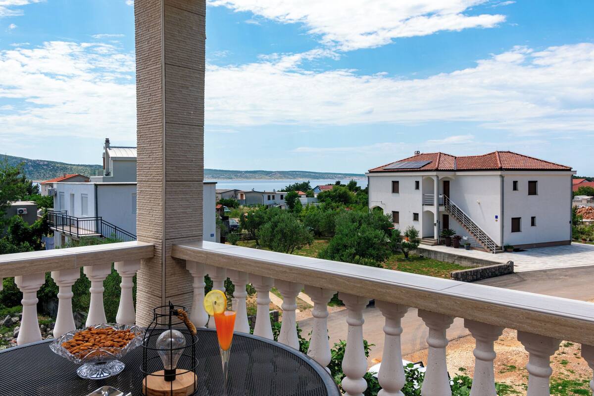 Ein Balkon mit Esstisch, Blick auf ein weißes Gebäude mit rotem Dach und dahinter ein Gewässer.