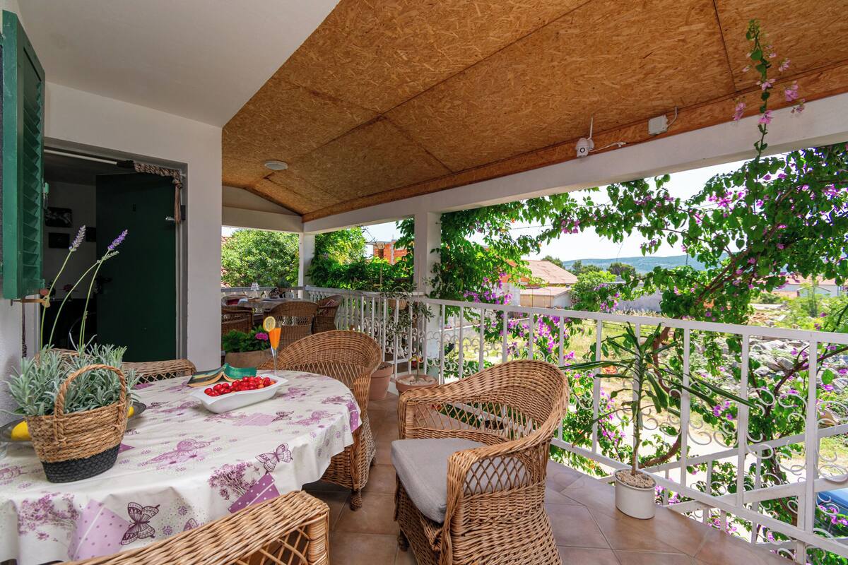 Eine überdachte Terrasse mit Korbmöbeln, einem Tisch mit einem Blumen-Tischläufer und Blumentöpfen.