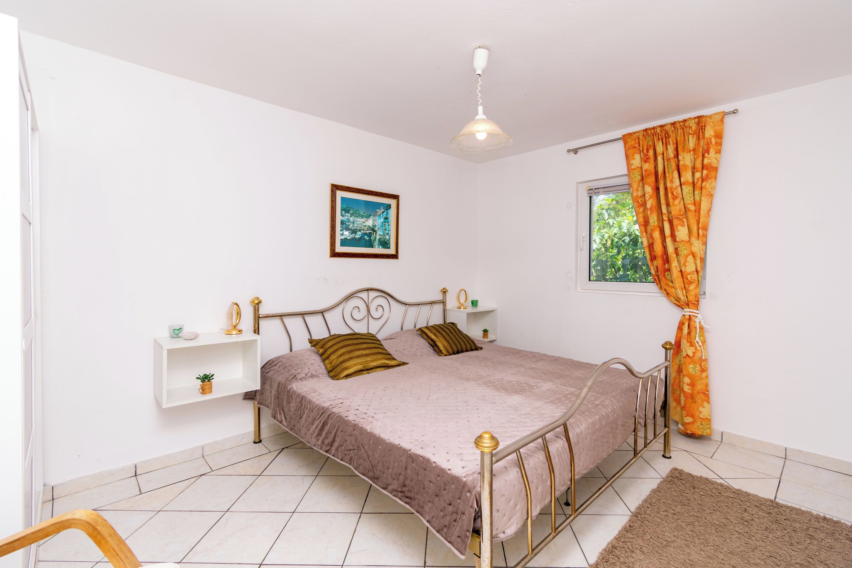 Ein Schlafzimmer mit Bett, Nachttisch, Ablagefläche, Fenster mit Vorhängen, einem Bild an der Wand und einer Deckenleuchte.