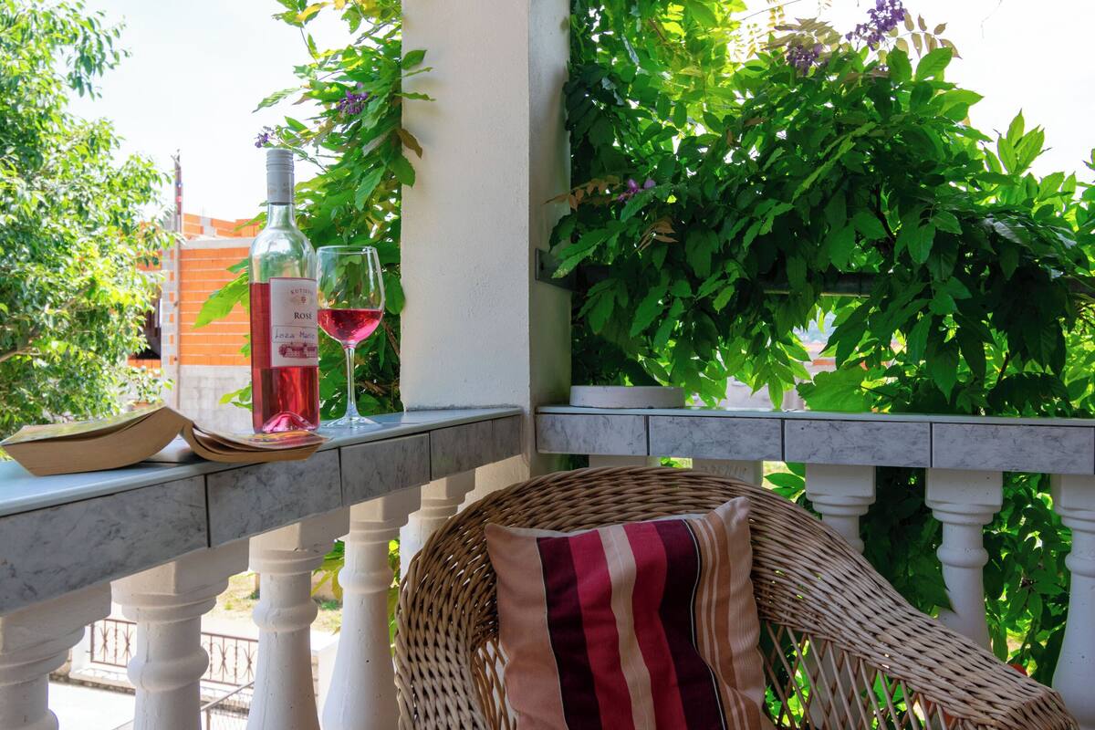 Ein Korbstuhl mit gestreiftem Kissen, eine Flasche Roséwein und ein Glas auf einem Balkongeländer.