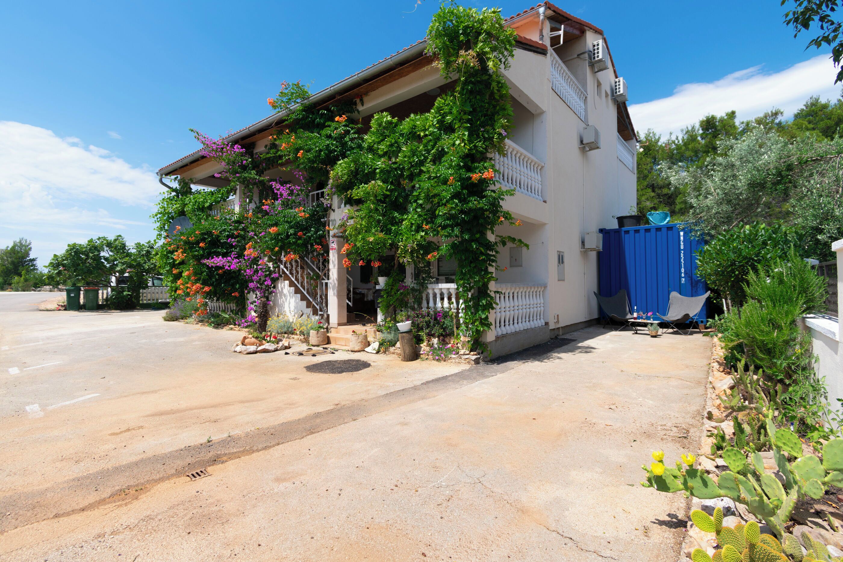 Un edificio de dos pisos con un balcón, un contenedor de basura azul y un jardín con cactus y plantas con flores.