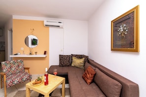 Una sala de estar con un sofá, una silla, una mesita con una botella y un vaso, un espejo y un cuadro en la pared.