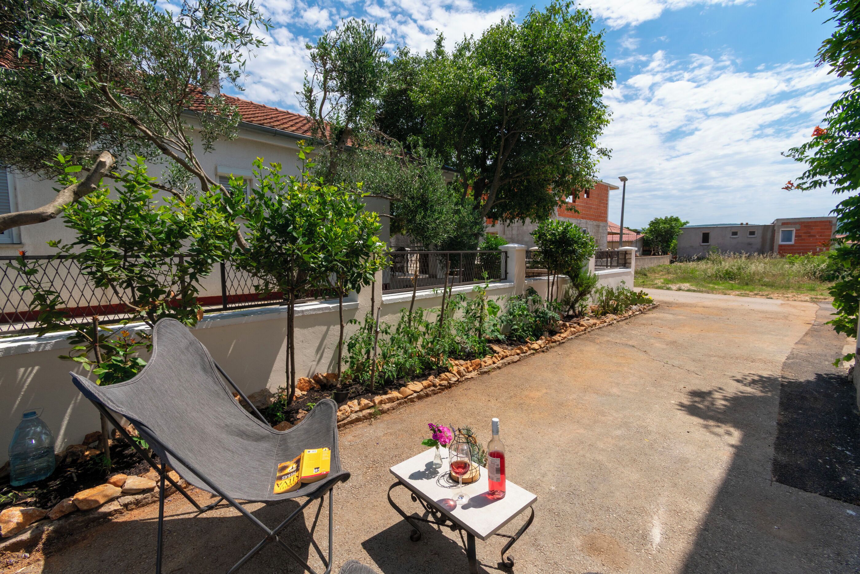 Un patio con una silla de metal, una mesa pequeña con una botella y flores, y una valla con árboles.