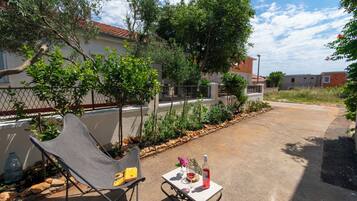 Un patio con una silla de metal, una mesa pequeña con una botella y flores, y una valla con árboles.