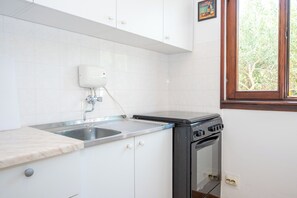 A kitchen with white cabinets, a stainless steel sink, a stove, and a window with a view of trees. - Apartments Olive and Sea (Solta)