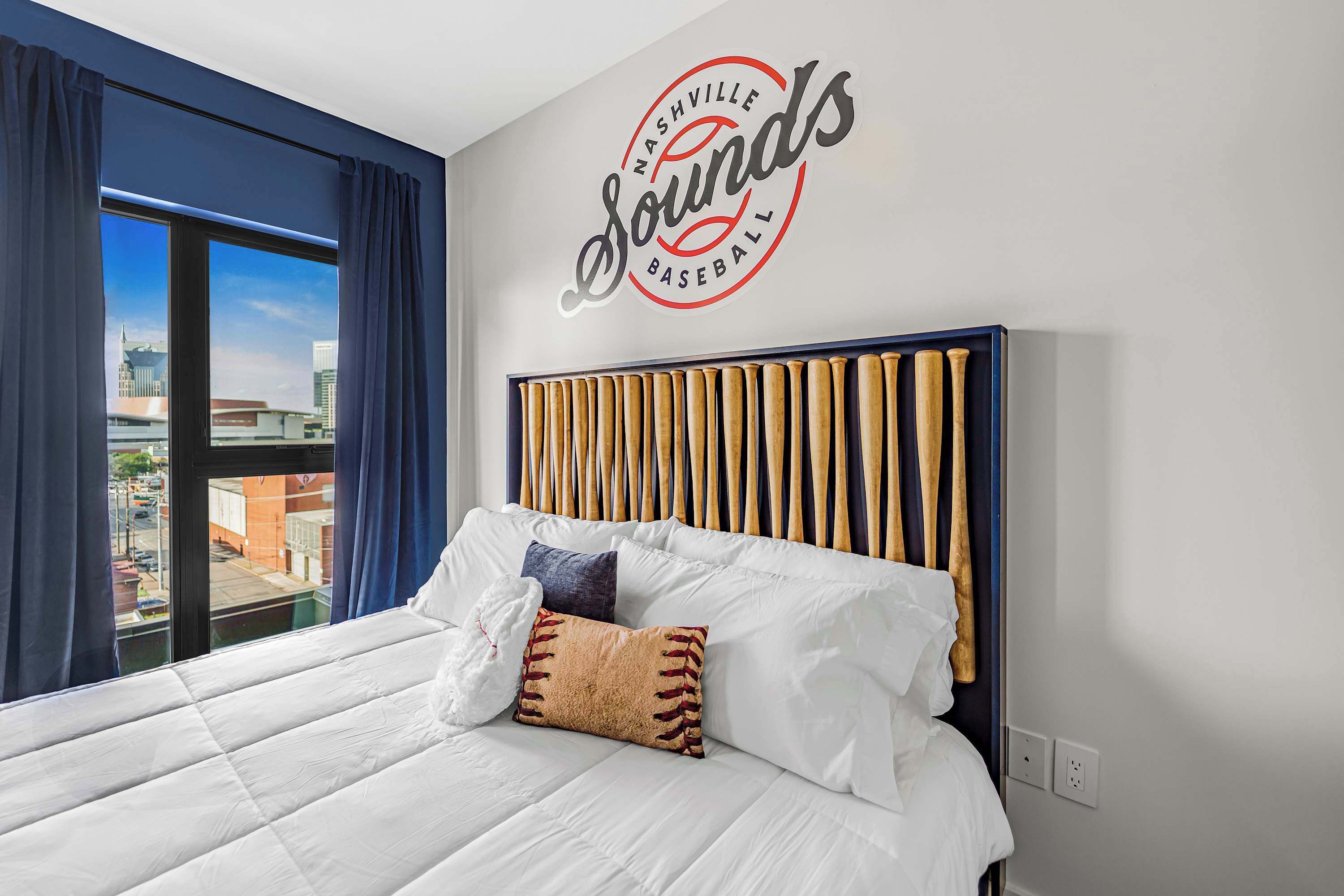 Quarto de hotel com cama, cabeceira com tacos de beisebol, janela com cortinas e um letreiro na parede que diz 'Nashville Sounds Baseball'.