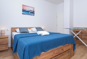 Ein Schlafzimmer mit einem hölzernen Bett, einer blauen Bettdecke, einem weißen Kissen, einem grauen Kissen, einem zusammengerollten weißen Handtuch, einem Nachttisch mit einer Lampe und einem Bild von Booten an der Wand.