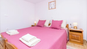 Een slaapkamer met een roze sprei, twee kussens, een houten nachtkastje met een lamp en een aan de muur hangend schilderij.