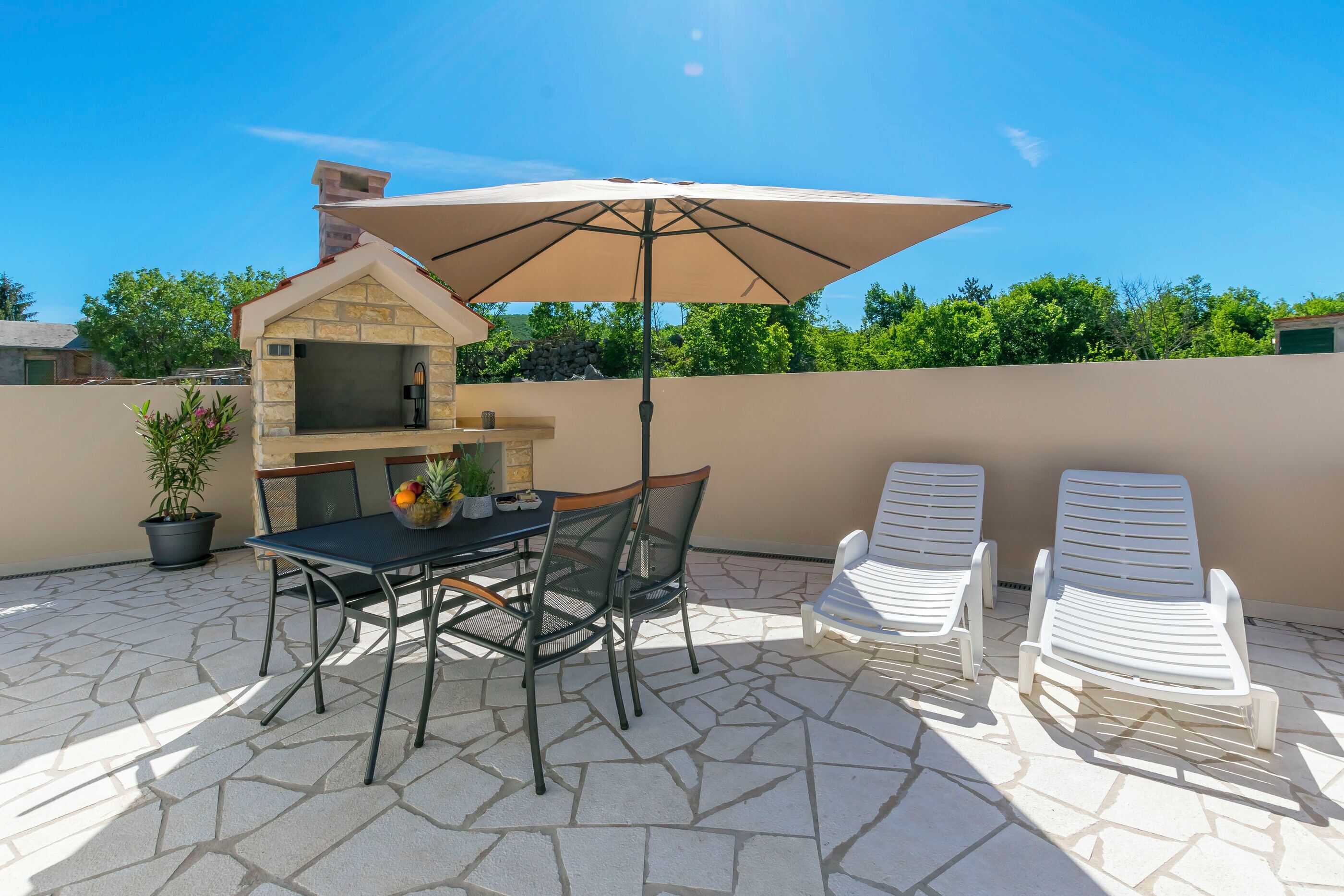 Une terrasse extérieure avec un sol en pierre, un barbecue, une table avec des chaises et deux fauteuils de jardin.