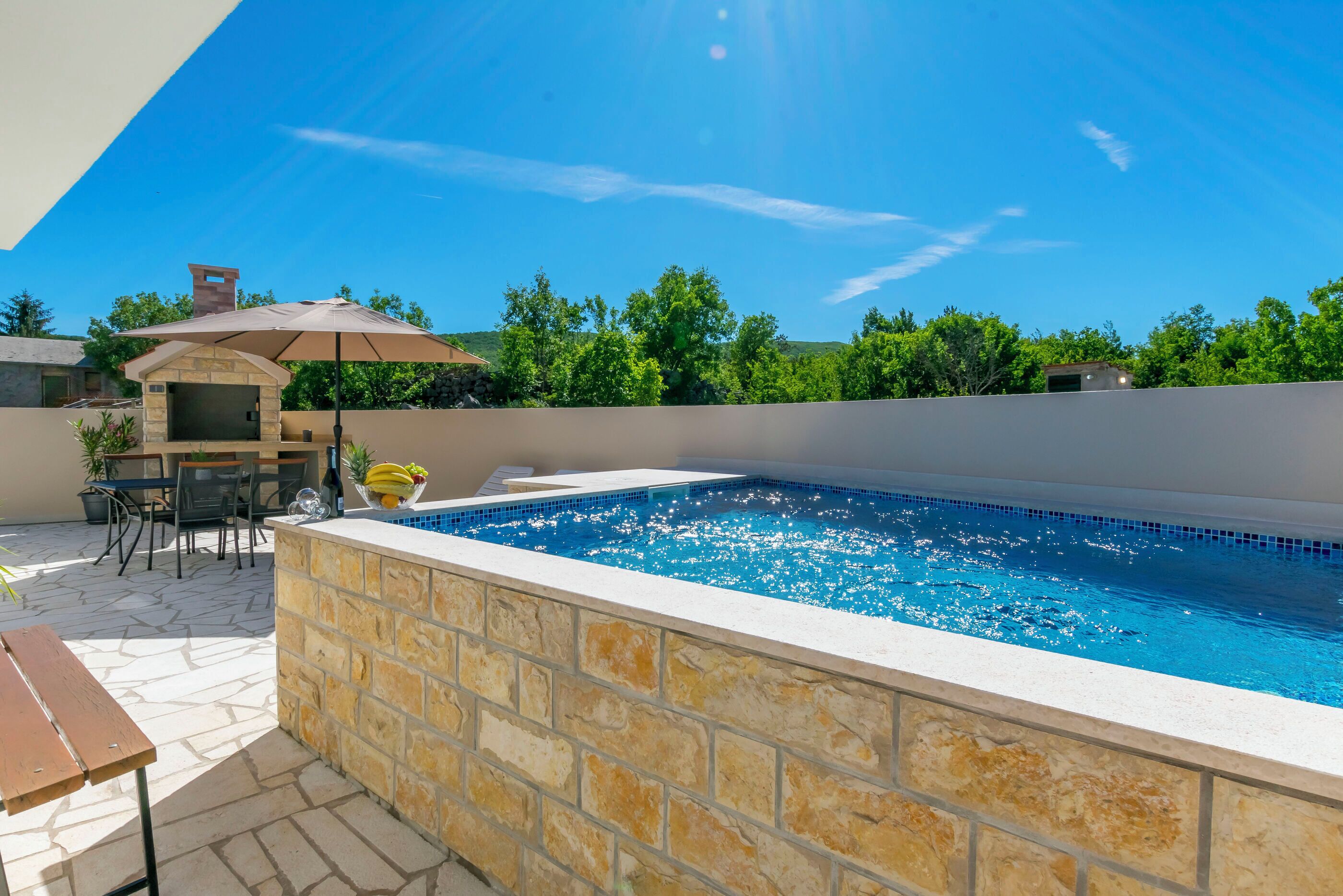 Une piscine avec des murs en pierre, un espace barbecue et une terrasse avec une table et des chaises.