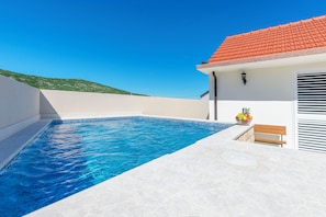 A swimming pool with clear blue water, a red-tiled roof, and a white exterior wall. - House Romance for two (Lovreć)