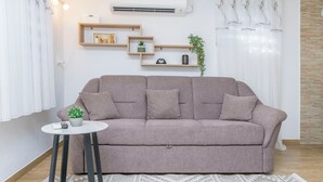 Een woonkamer met een bank, een klein bijzettafeltje, een wandplank met decoratieve objecten en een raam met gordijnen.
