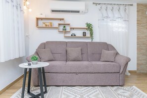 Ein Wohnzimmer mit einer Couch, einem kleinen Beistelltisch, einem Wandregal mit Dekoration und einem Fenster mit Vorhängen.