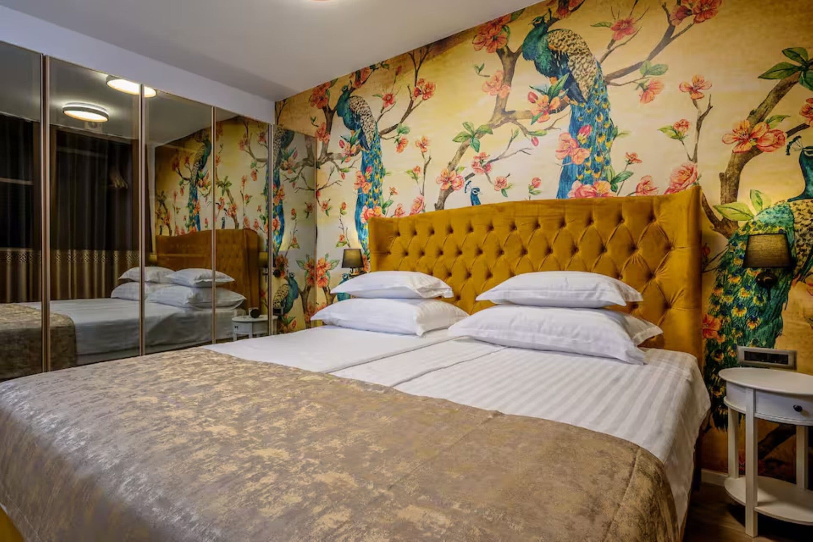 Une chambre d’hôtel avec un grand lit, un miroir, un mur orné d’une fresque représentant un paon et des motifs floraux, et une petite table de chevet.