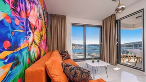 A modern living room with a colorful abstract mural, a plush orange sofa, a small white table, and a view of the sea through large glass doors. - Apartments Penthouse Lux (Rogoznica)