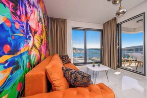 A modern living room with a colorful abstract mural, a plush orange sofa, a small white table, and a view of the sea through large glass doors. - Apartments Penthouse Lux (Rogoznica)