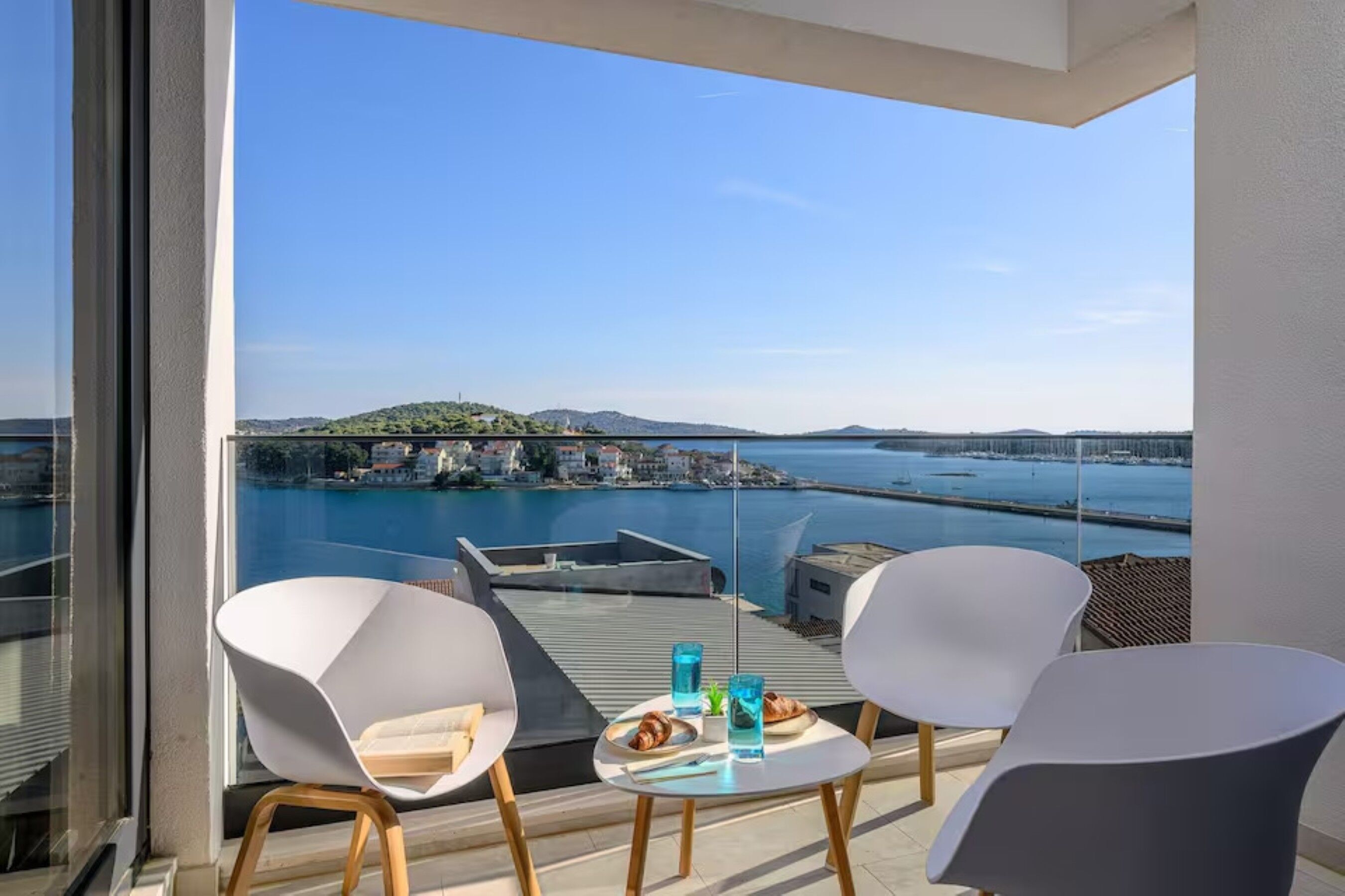 Ein Balkon mit zwei weißen Stühlen, einem kleinen Tisch und Blick auf ein Gewässer sowie entferntes Land.