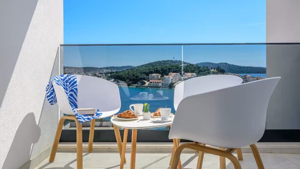A balcony with two white chairs, a small table, and a view of a coastal area with hills and buildings. - Apartments Penthouse Lux (Rogoznica)